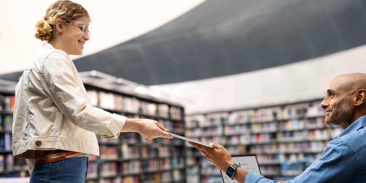 : Ein Lehrer überreicht einer Schülerin in einer Bibliothek ein iPad, weiter Geräte liegen auf dem Tisch vor ihm