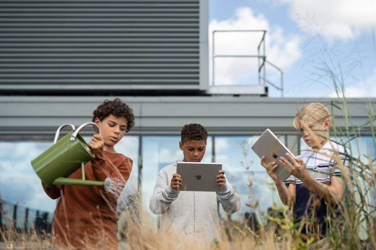 Zwei Schüler arbeiten mit dem iPad in einem begrünten Gelände, ein dritter benutzt eine Gießkanne