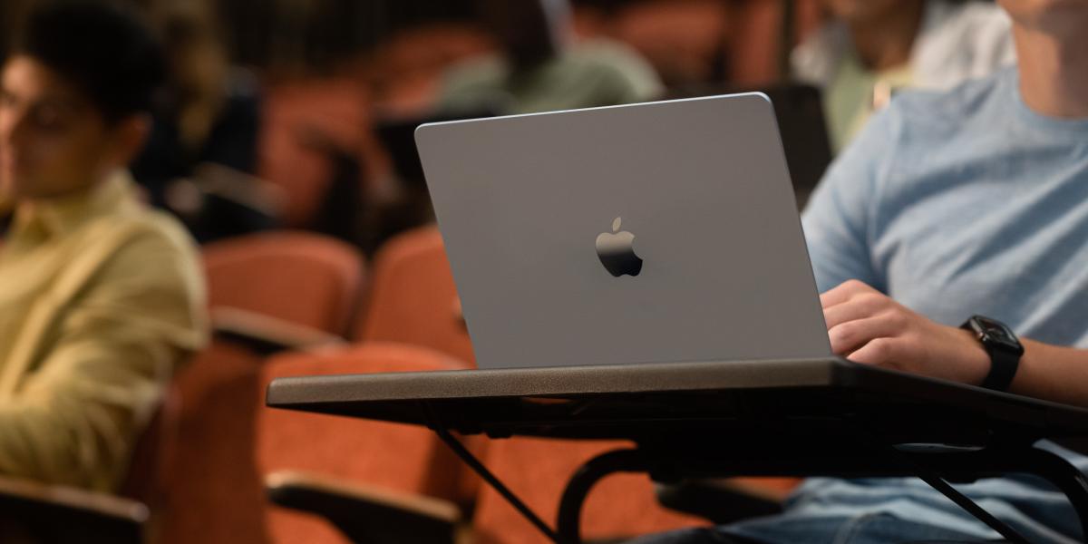 Ein Student verfolgt in einem Uni Auditorium einen Vortrag, vor ihm ein aufgeklapptes MacBook
