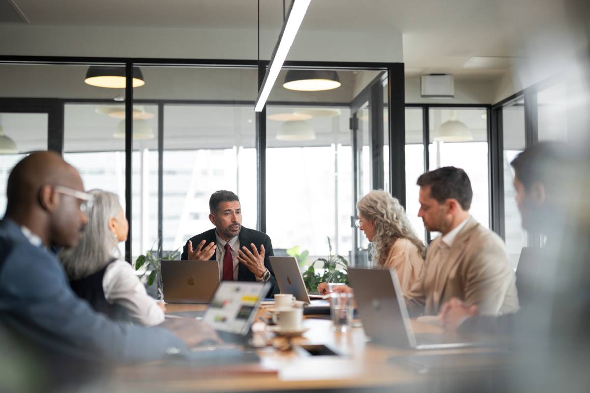 Sechs Personen bei einem Meeting in einer Bildungseinrichtung, am Tisch stehen mehrere MacBooks